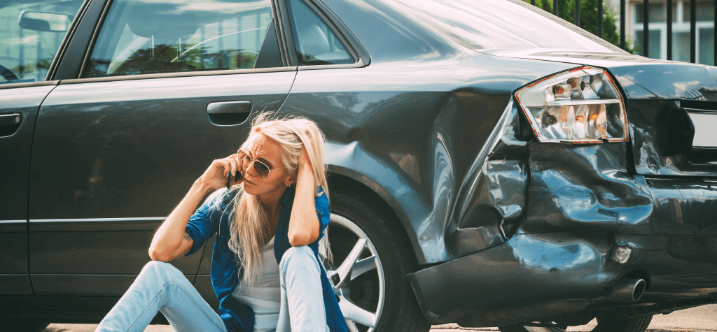 A person sits against a car that is crushed from a car accident