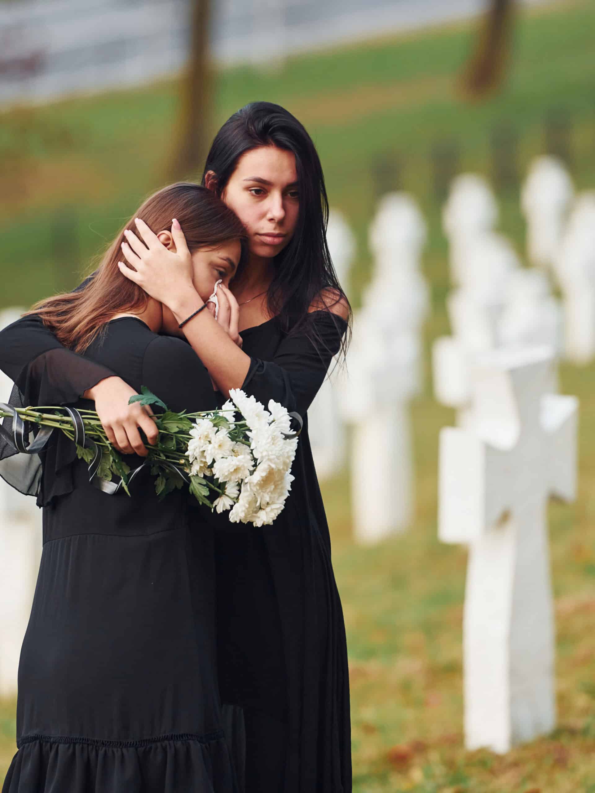 Embracing each other and crying. Two young women in black clothes visiting cemetery with many white crosses. Conception of funeral and death