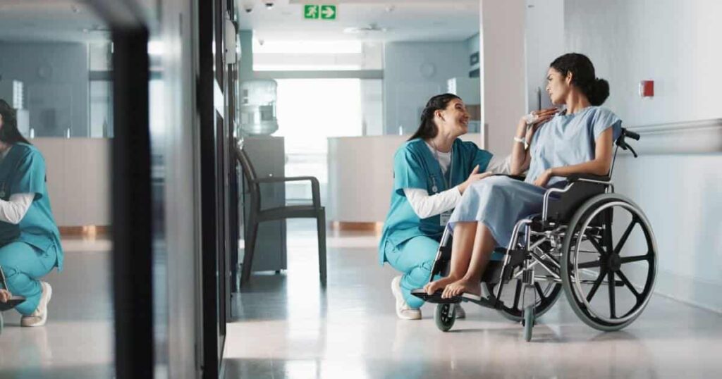 A nurse kneels next to a woman wearing a hospital gown sitting in a wheelchair in a hospital hallway