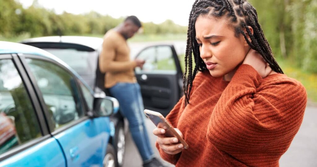 Woman holding her neck in pain, looking at phone near a car accident scene with another person in the background.