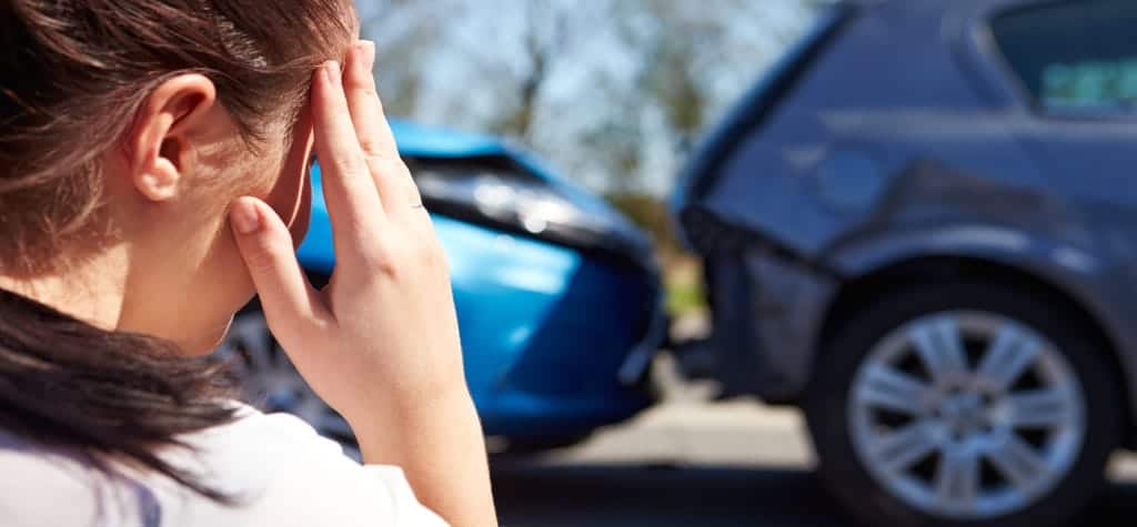 A woman holding her head in distress after a car accident, with two vehicles damaged in the background.