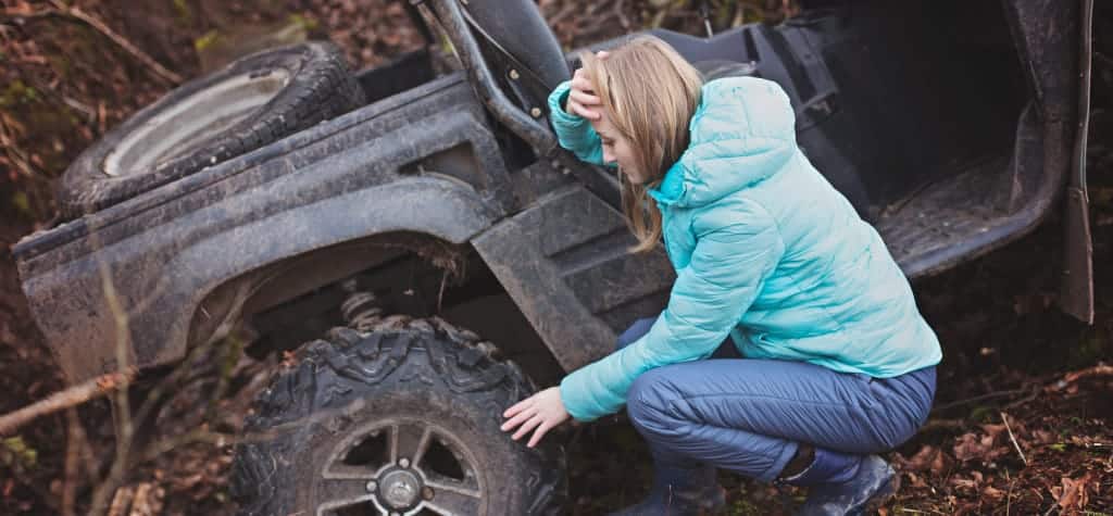 Women in a blue jacket inspecting a stuck ATV in a muddy forest area, looking concerned.