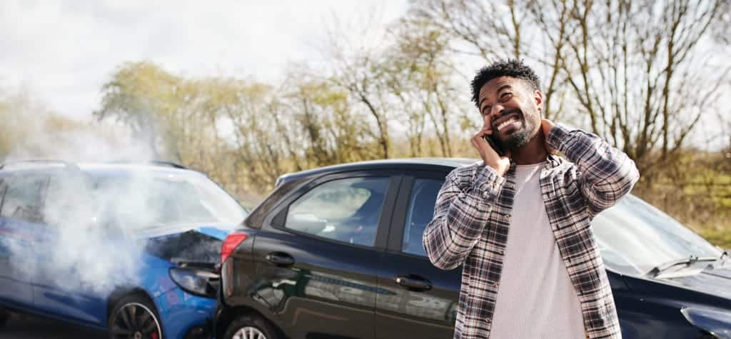 A man holding his neck and talking on the phone after a car accident, with two damaged vehicles in the background.